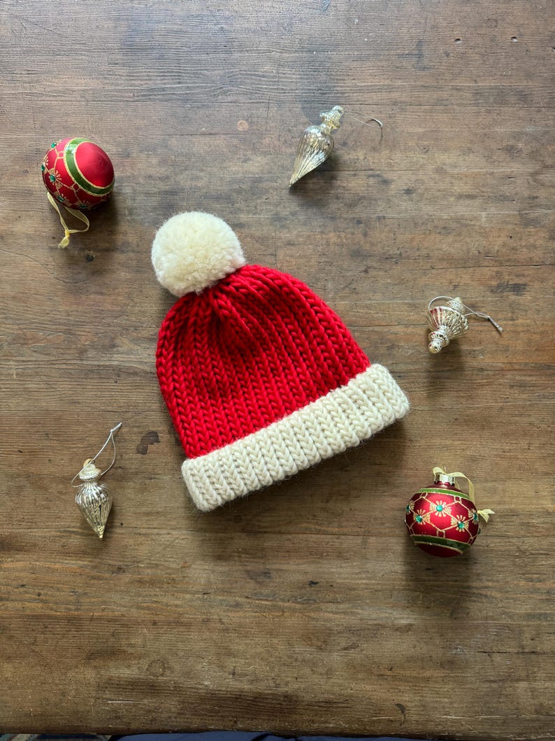 May include: A bright red knit beanie with a thick, cream-colored ribbed cuff and a matching cream-colored pom-pom on top. The hat is surrounded by two round red and green patterned ornaments and two silver-toned teardrop-shaped ornaments.