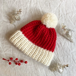 May include: A chunky knit beanie featuring a vibrant red main section and a thick cream-colored ribbed brim. The hat is topped with a large, fluffy cream-colored pom-pom. The knit pattern is vertical and textured, showcasing a classic winter style.