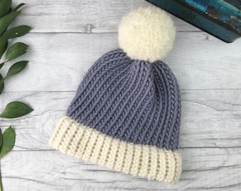 Puede incluir: Un gorro de punto con un pompón blanco en la parte superior. El gorro es de dos tonos, con la parte superior gris y el borde blanco.