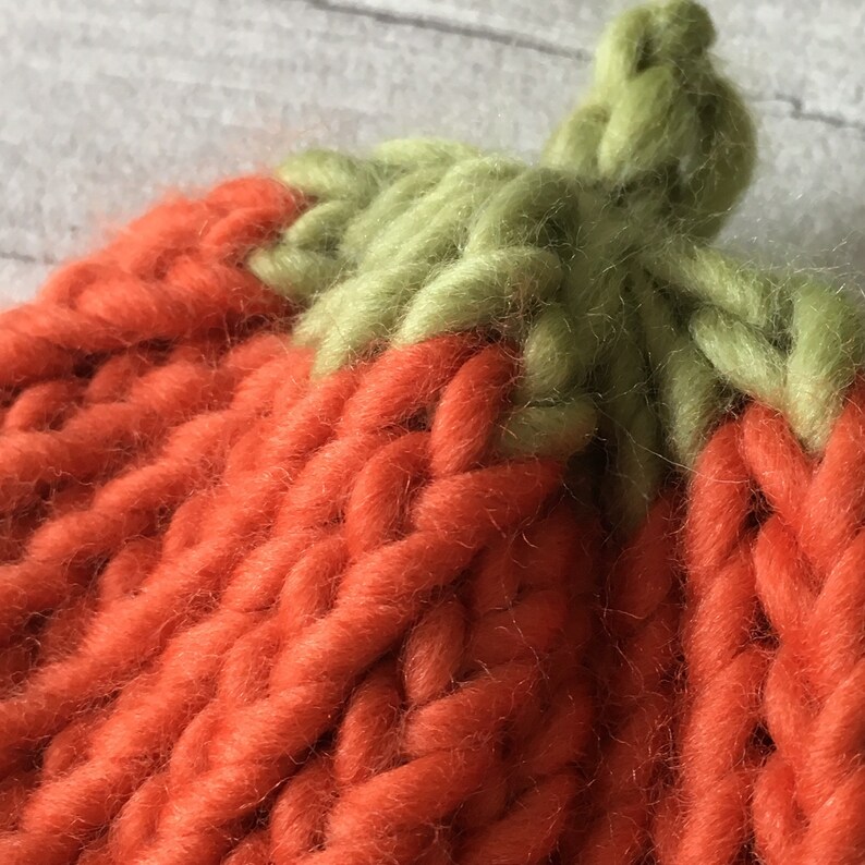 Puede incluir: Primer plano de un gorro de punto de calabaza con un tallo verde. El gorro est&aacute; hecho de hilo naranja y tiene un punto texturizado.