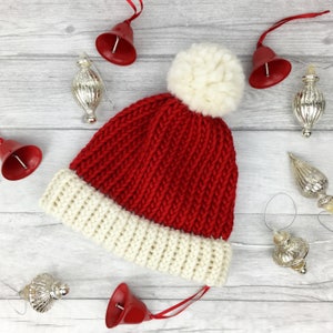 Hand santa knitted hat with cream brim, red middle and a fluffy cream pom pom. Hat shown is adult size but available in range of sizes. The hat is surrounded by Christmas baubles as props. The hat is cosy and warm looking with thick wool