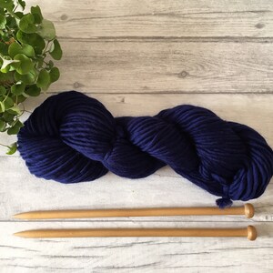 Puede incluir: Un ovillo de lana azul oscuro con dos agujas de tejer de madera.