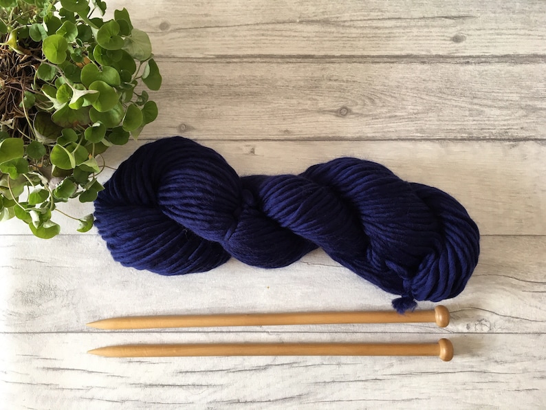 Puede incluir: Un ovillo de lana azul oscuro con dos agujas de tejer de madera.