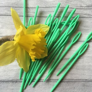 Puede incluir: Un montón de agujas de plástico verde para lana, también conocidas como agujas de tapicería, están dispuestas en forma de abanico. Una flor de narciso amarilla está en primer plano.