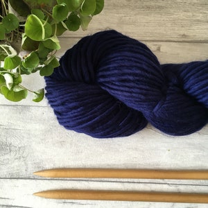 Puede incluir: Un ovillo de lana azul oscuro con dos agujas de tejer de madera.