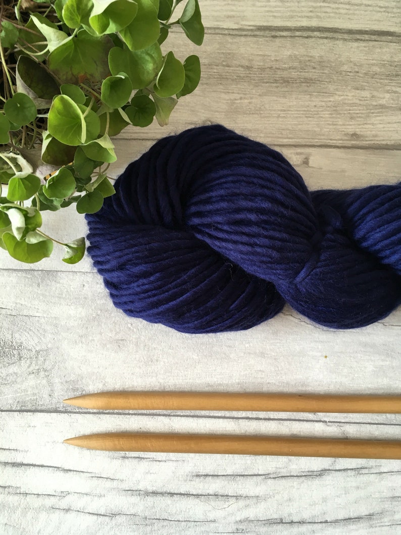 Puede incluir: Un ovillo de lana azul oscuro con dos agujas de tejer de madera.