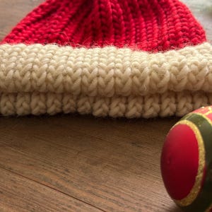 May include: A hand-knitted beanie with a vibrant red crown and a thick, cream-colored ribbed cuff. The hat is topped with a fluffy white pom-pom. Beside the beanie sits a round, red and green decorative ornament with gold-colored accents.