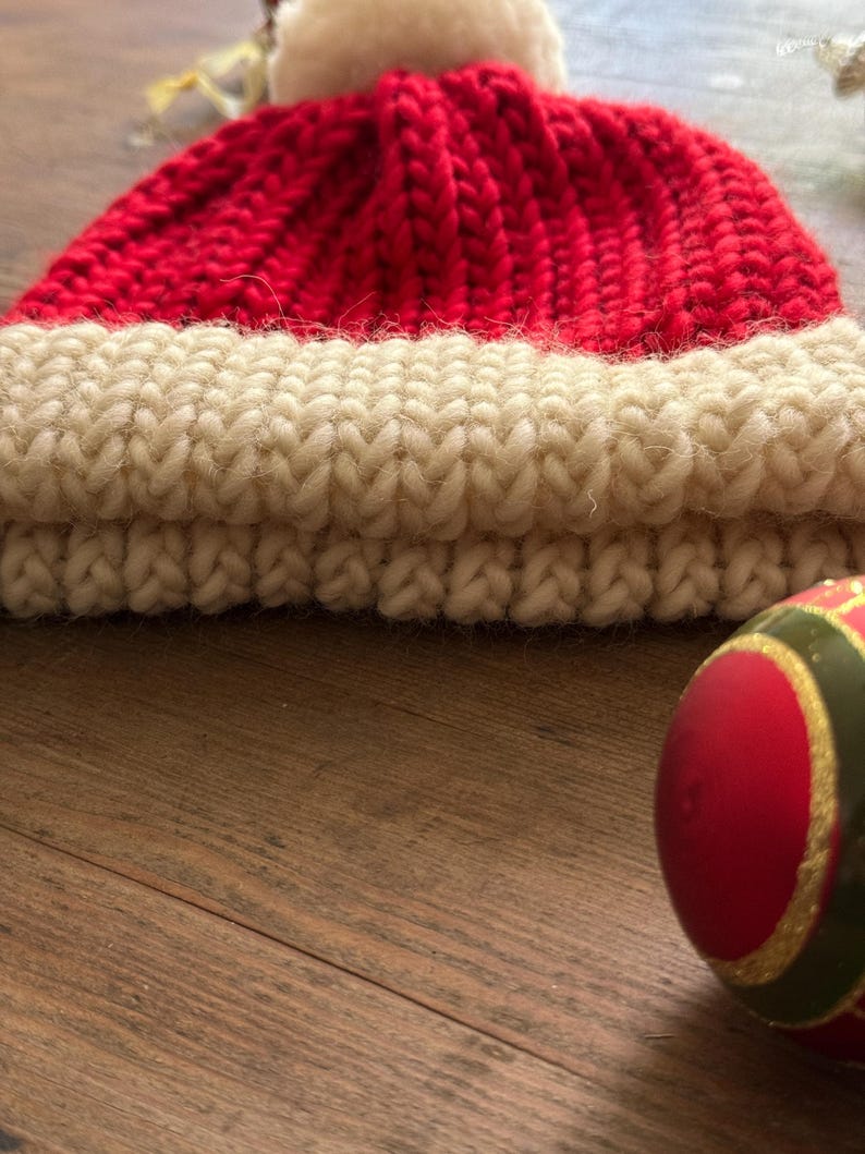 May include: A hand-knitted beanie with a vibrant red crown and a thick, cream-colored ribbed cuff. The hat is topped with a fluffy white pom-pom. Beside the beanie sits a round, red and green decorative ornament with gold-colored accents.
