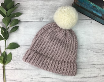 Puede incluir: Un gorro de punto color malva con un pompón blanco en la parte superior.