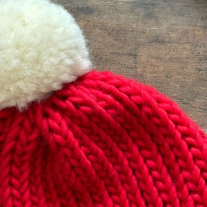 May include: A bright red knit beanie featuring a thick, chunky yarn texture and a large, fluffy off-white pom-pom attached to the top. The hat is constructed with a vertical ribbed stitch pattern, showcasing the vibrant color and soft material.