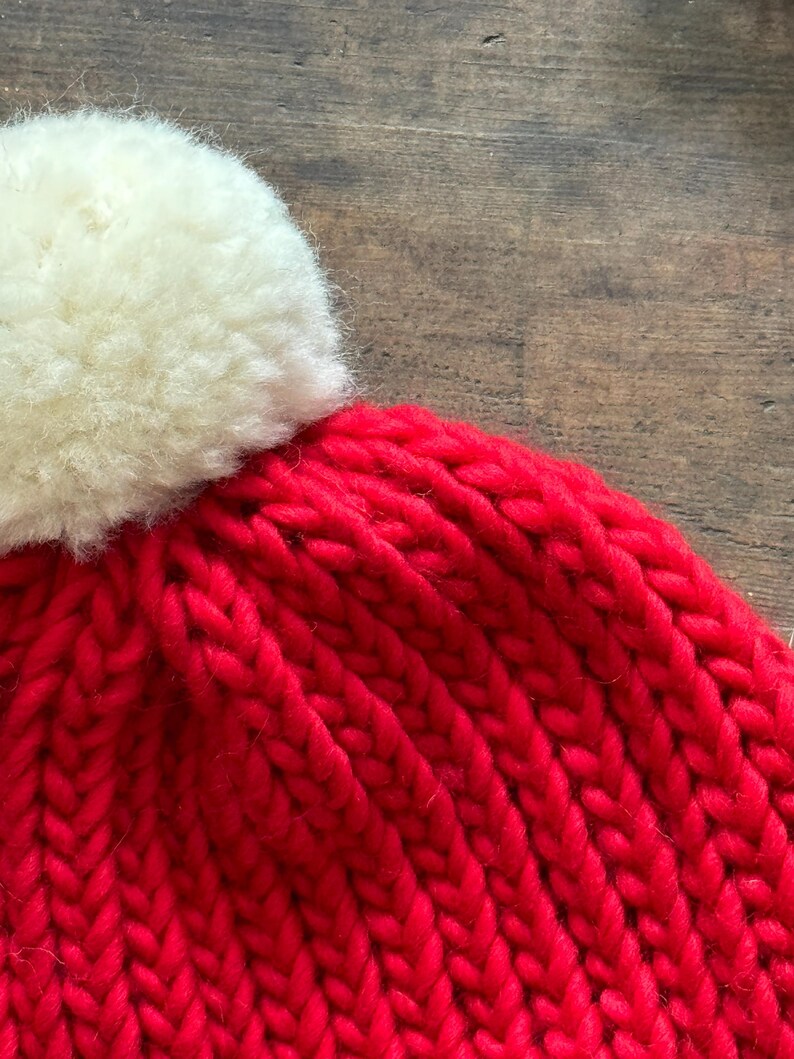 May include: A bright red knit beanie featuring a thick, chunky yarn texture and a large, fluffy off-white pom-pom attached to the top. The hat is constructed with a vertical ribbed stitch pattern, showcasing the vibrant color and soft material.