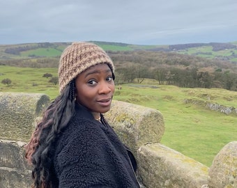 Puede incluir: Una persona con un gorro de punto marrón y beige y una chaqueta de vellón negra se encuentra de pie en un muro de piedra con vistas a un campo verde y un bosque en la distancia.