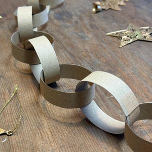 May include: A paper chain made from beige card, with interlocking links. Gold Christmas decorations, including a star and a bell, are scattered on a wooden surface. The image evokes a festive, DIY holiday craft project, perfect for the Christmas season.