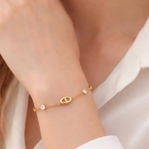 Peut inclure: Un bracelet doré avec un pendentif ovale central et trois petites pierres précieuses rondes et transparentes. Le bracelet est délicat et composé de fines maillons de chaîne. Porté sur un poignet, contre une chemise blanche.
