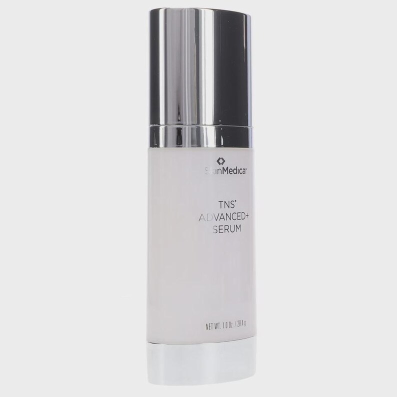 May include: A cylindrical SkinMedica TNS ADVANCED+ SERUM bottle with a metallic silver cap and base. The white opaque container features black text reading SkinMedica, TNS ADVANCED+ SERUM, and NET WT. 1.0 OZ / 28.4 G printed on the front surface.