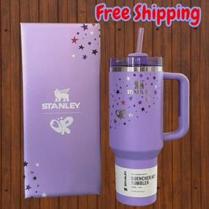 May include: A light purple Stanley Quencher H2.0 tumbler with a matching handle, lid, and straw. The tumbler features a silver star pattern and the Stanley logo. It is displayed next to a matching light purple box with the Stanley logo and stars.