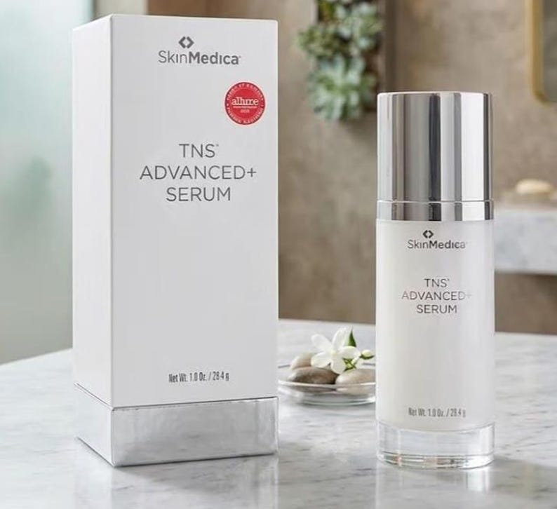 May include: A white SkinMedica TNS ADVANCED+ SERUM box and a matching cylindrical pump bottle. The packaging features black text, a red circular award seal, and states a net weight of 1.0 oz. / 28.4 g. The bottle has a metallic silver cap.