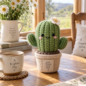 Puede incluir: Un cactus de ganchillo verde con una pequeña flor blanca en la parte superior, ojos negros y mejillas rosadas, sentado en una maceta de ganchillo beige. La maceta tiene una etiqueta de tela rectangular con el texto Good Things Grow Here y un corazón.