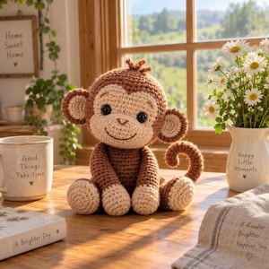 Può includere: Un giocattolo a forma di scimmia all'uncinetto marrone e beige con grandi orecchie e coda arricciata, seduto su una superficie di legno accanto a una tazza in ceramica bianca con il testo Good Things Take Time e un vaso bianco con margherite e il testo Happy Little Moments.
