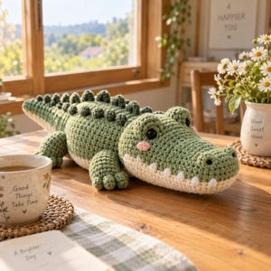 May include: A sage green crocheted alligator plush toy with a textured back, dark eyes, and pink cheeks. The alligator has a cream-coloured underbelly and a row of small white teeth. It is positioned on a wooden surface next to a ceramic mug.