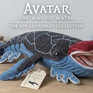 Könnte beinhalten: Gehäkeltes Wal-Spielzeug aus der Avatar: The Way of Water Sea Guardians Collection. Das Spielzeug hat einen grauen Körper, blaue Flossen und einen roten Kopf. Der Wal hat ein offenes Maul mit rosa Details und ein Etikett mit "crochet Tuikuri".