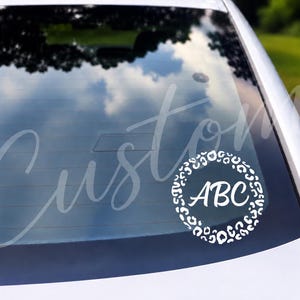 May include: A white circular decal applied to a car rear window, featuring a leopard print border surrounding the letters ABC in a script font. The design is solid white and contrasts against the dark glass of the vehicle's rear windshield.