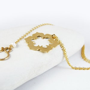 May include: A gold necklace with a delicate chain and a large, openwork pendant in the shape of a flower. The pendant has a cutout design and is attached to the chain with a small loop. A single pearl bead is visible on the chain.