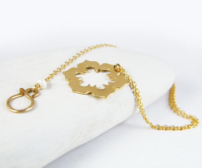 May include: A gold necklace with a delicate chain and a large, openwork pendant in the shape of a flower. The pendant has a cutout design and is attached to the chain with a small loop. A single pearl bead is visible on the chain.