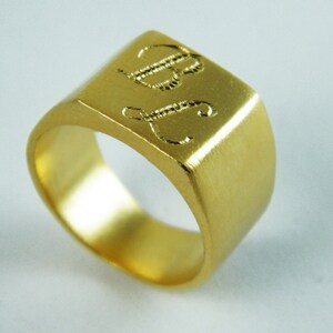 May include: A gold-colored signet ring with a square face. The ring features engraved initials "B" and "J" on the top surface. The ring has a brushed metal finish and a rounded interior band.
