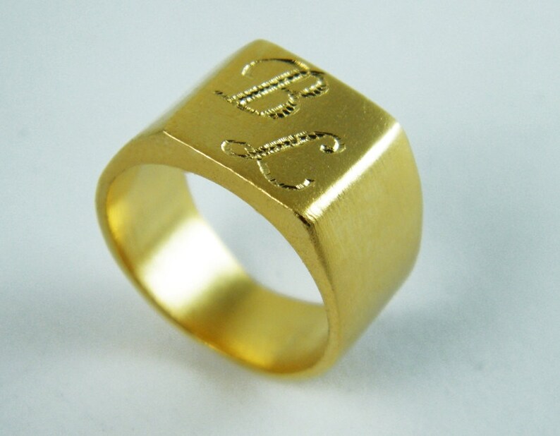May include: A gold-colored signet ring with a square face. The ring features engraved initials "B" and "J" on the top surface. The ring has a brushed metal finish and a rounded interior band.