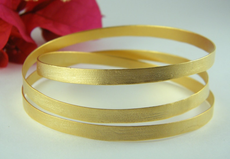 May include: Three gold-toned bangle bracelets stacked on top of each other. The bracelets have a brushed finish.