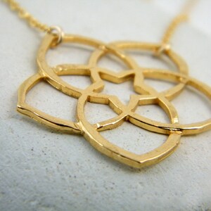 May include: A gold-toned necklace with a delicate, geometric pendant featuring a flower-like design. The pendant is made of intricate, interwoven circles and has a slightly hammered texture.