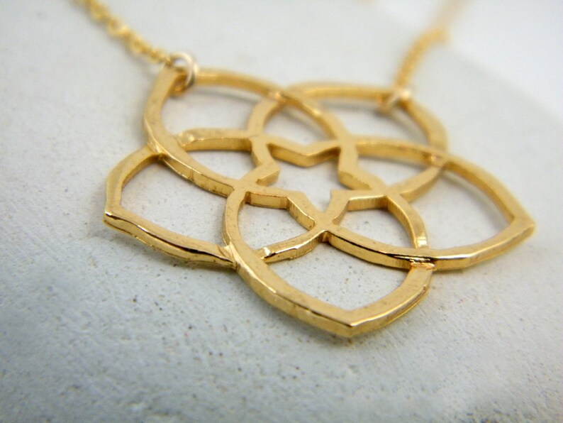 May include: A gold-toned necklace with a delicate, geometric pendant featuring a flower-like design. The pendant is made of intricate, interwoven circles and has a slightly hammered texture.