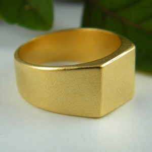 May include: A gold-colored ring with a rectangular face and a thick band. The ring has a matte finish and a simple, modern design. The ring is a signet style and is likely made of metal.