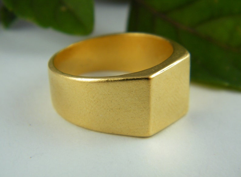 May include: A gold-colored ring with a rectangular face and a thick band. The ring has a matte finish and a simple, modern design. The ring is a signet style and is likely made of metal.