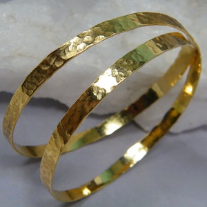 Puede incluir: Dos pulseras de brazalete de oro martilladas. Las pulseras están apiladas una encima de la otra y están hechas de un metal grueso y texturizado.