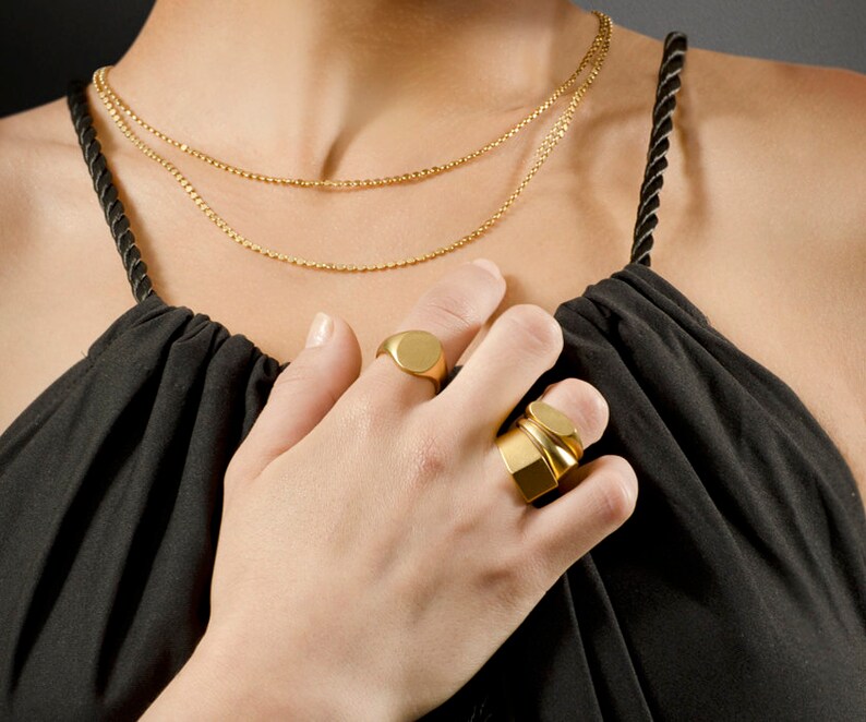 May include: Gold-toned jewelry set featuring two delicate chain necklaces and two rings. One ring is a classic signet style, and the other is a multi-layered design. The jewelry is displayed against a black top.