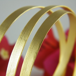 May include: Three gold-colored metal bangles stacked on top of each other. The bangles are smooth and have a brushed finish.