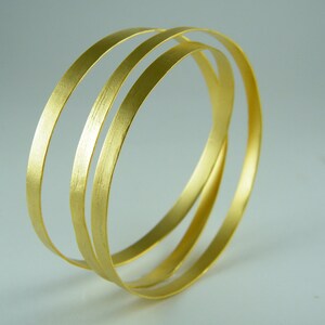 May include: Three gold-colored metal bangles stacked on top of each other. The bangles are smooth and shiny.
