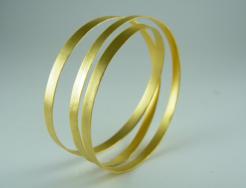 May include: Three gold-colored metal bangles stacked on top of each other. The bangles are smooth and shiny.