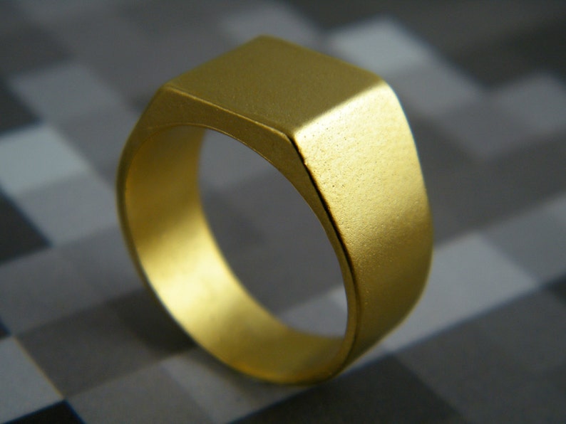 May include: A gold-colored signet ring with a square face. The ring has a simple, classic design and a matte finish. The ring is made of metal and is likely worn on a finger.