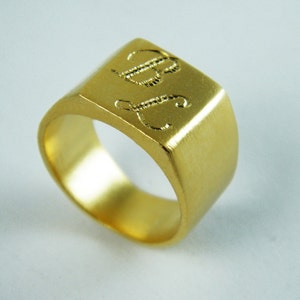 May include: A gold-colored signet ring with a rectangular face. The face of the ring is engraved with the initials "BJ" in a cursive font. The ring has a brushed finish and a thick band.