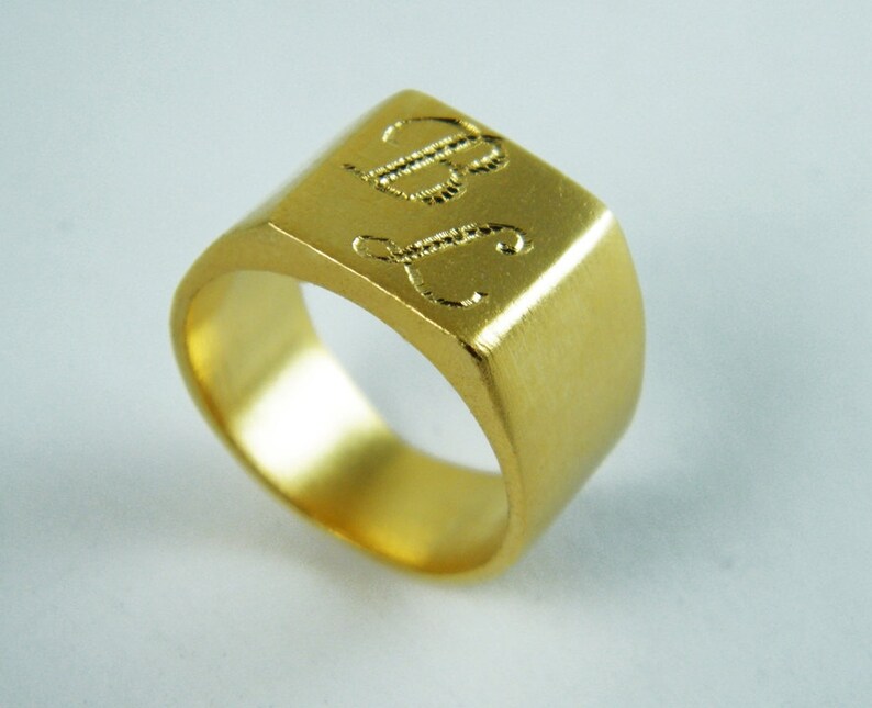 May include: A gold-colored signet ring with a rectangular face. The face of the ring is engraved with the initials "BJ" in a cursive font. The ring has a brushed finish and a thick band.
