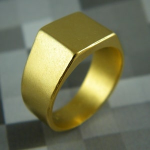 May include: A gold-colored signet ring with a square face. The ring has a smooth, matte finish and a classic, minimalist design. The ring is a solid band of gold.