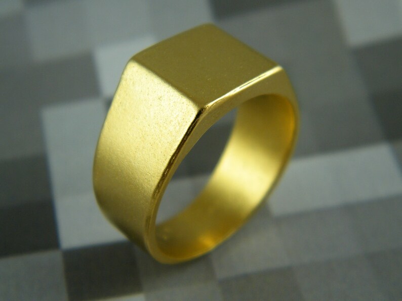 May include: A gold-colored signet ring with a square face. The ring has a smooth, matte finish and a classic, minimalist design. The ring is a solid band of gold.