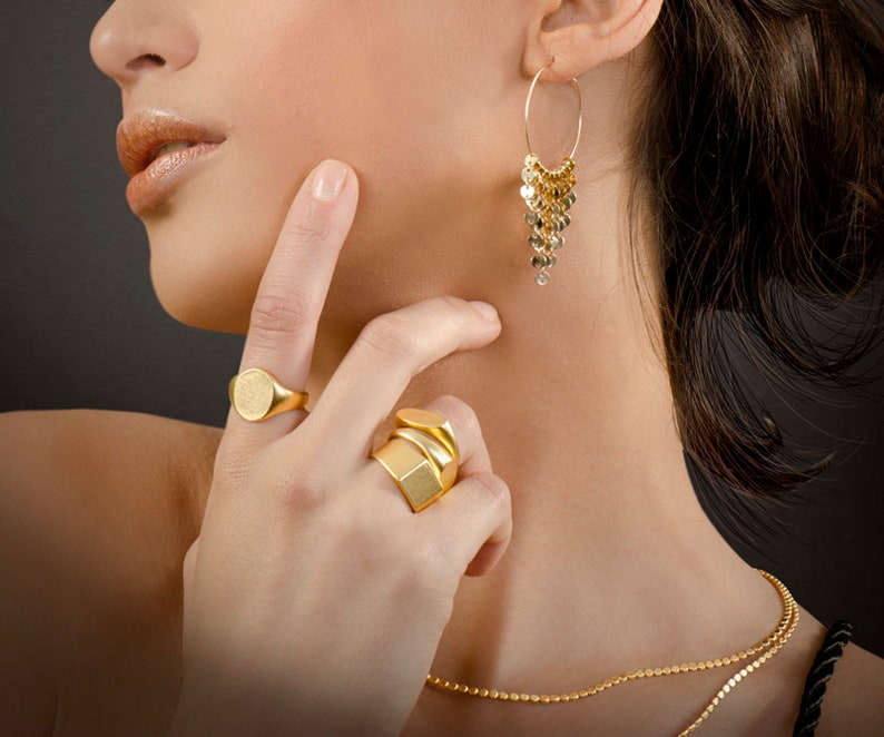 Puede incluir: Una mujer lleva joyas de oro, incluyendo un par de pendientes de aro con una cadena colgante de c&iacute;rculos dorados, un anillo de oro con una cara redonda y un anillo de oro con un dise&ntilde;o geom&eacute;trico. Tambi&eacute;n lleva un collar de cadena de oro.