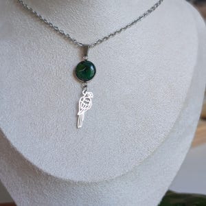 May include: A silver-toned necklace featuring a pendant with a round, dark green stone and a parrot-shaped charm. The necklace is displayed on a white mannequin, showcasing the jewelry's design and details. The chain is delicate and the parrot charm is a simple outline.