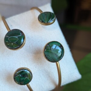 May include: Four gold-toned bracelets with circular green stone accents. The stones have a marbled appearance with shades of green and black. The bracelets are displayed on a white surface, showcasing their design and color.