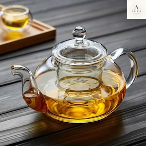 Può includere: Una teiera in vetro trasparente con coperchio abbinato e un infusore interno in vetro. La teiera è riempita con un liquido color oro e presenta un manico curvo e un beccuccio corto. Una piccola tazza in vetro è visibile sullo sfondo.
