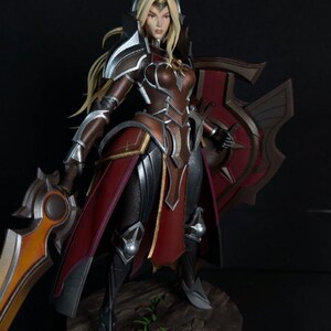 May include: A detailed figurine of a female warrior in armor, holding a sword and shield. The figure is predominantly brown, red, and silver, with blonde hair. The base is a circular platform with a textured, stone-like appearance.